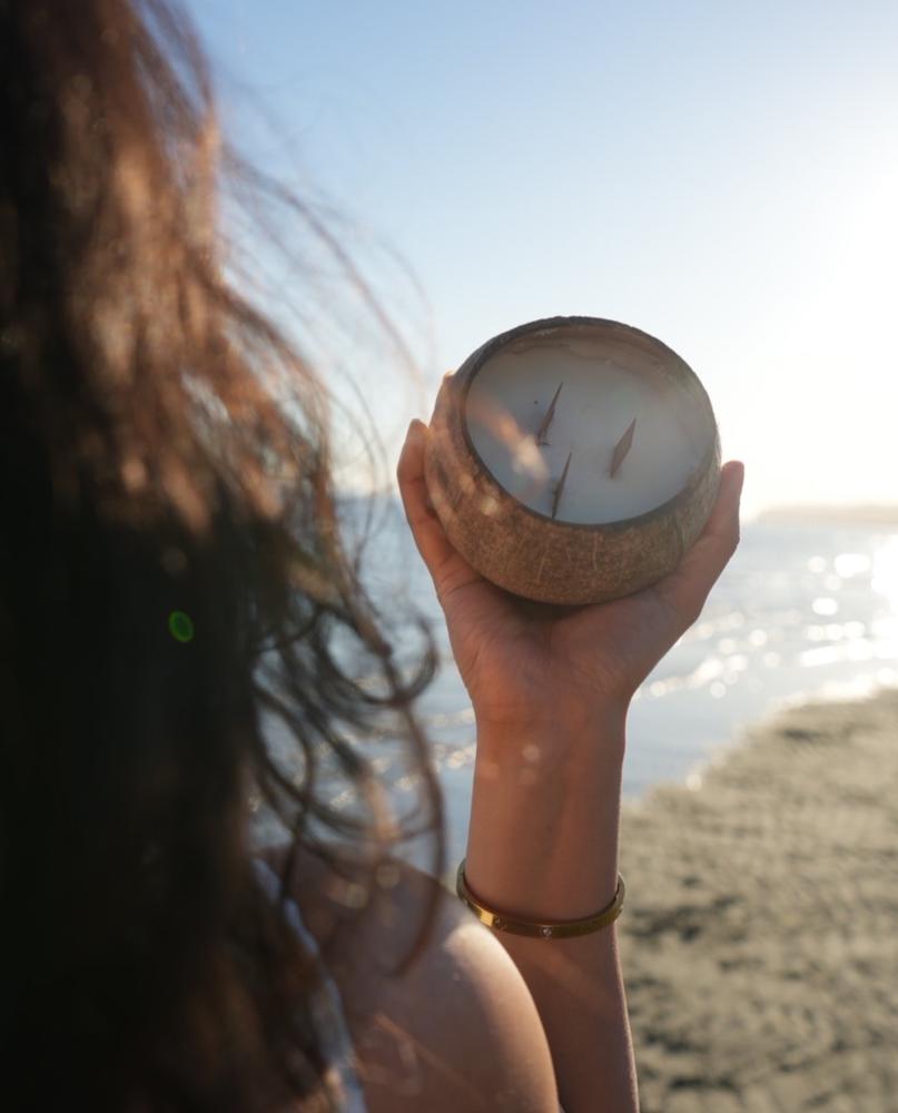Hand holding a coconut-shell candle aloft at the beach with sunlit ocean behind